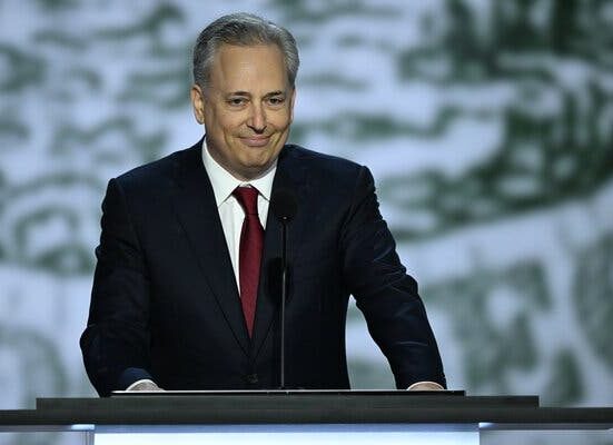 David Sacks, in a dark suit and red tie, standing behind a lectern with a microphone in front of him.