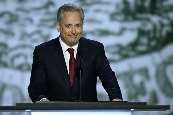 David Sacks, in a dark suit and red tie, standing behind a lectern with a microphone in front of him.